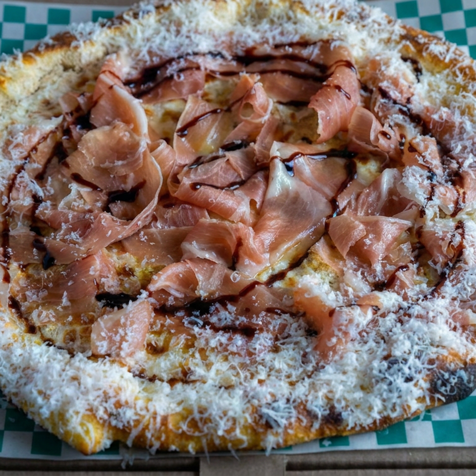Neapolitan-style pizza topped with prosciutto, melted cheese, and dark balsamic drizzle on checkered paper